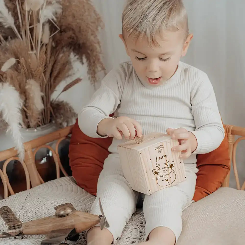Kind sitzt im Kinderzimmer und wirft MĂĽnze in personalisierte Holz-Spardose mit Reh-Motiv und eigenem Namen
