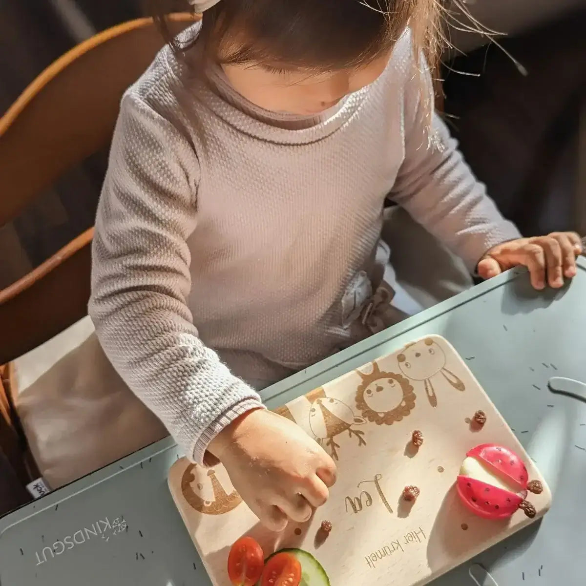 Kleinkind mit personalisiertem FrĂĽhstĂĽcksbrettchen beim Essen - niedliches Kind in hellem Langarmshirt verwendet ein Holzbrettchen mit Tiermotiven fĂĽr sein gesundes FrĂĽhstĂĽck mit GemĂĽse