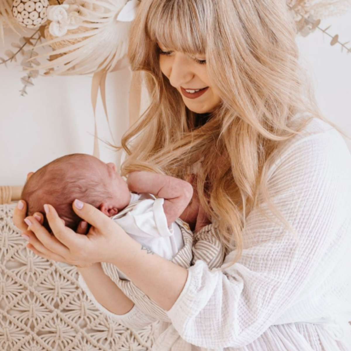 Junge Mutter mit langen blonden Haaren in weißem Kleid hält liebevoll ihr neugeborenes Baby im Arm. Sie sitzt auf einer gemütlichen Rattanbank mit gestrickten Kissen in Naturtönen. Im Hintergrund hängt ein dezenter Trockenblumenkranz in neutralen Farben an der weißen Wand. Die Szene strahlt Wärme, Geborgenheit und den besonderen Moment der frühen Mutterschaft aus."
