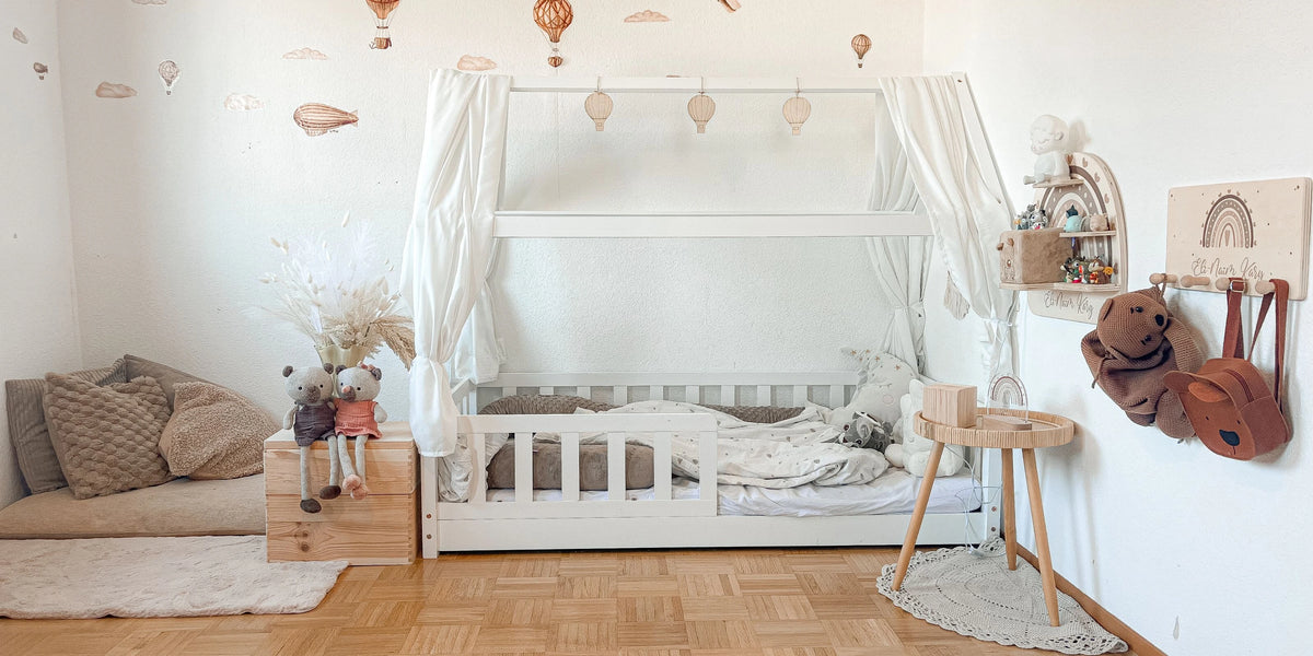Gemütliches Kinderzimmer im Montessori-Stil mit weißem Hausbett, warmen Holzelementen und dekorativer Tapete mit Heißluftballons und Luftschiffen. Der helle Raum ist liebevoll mit Stofftieren, einem Beistelltisch und Wanddekoration eingerichtet - perfekt für erholsamen Schlaf und kreatives Spielen in kindgerechter Atmosphäre.
