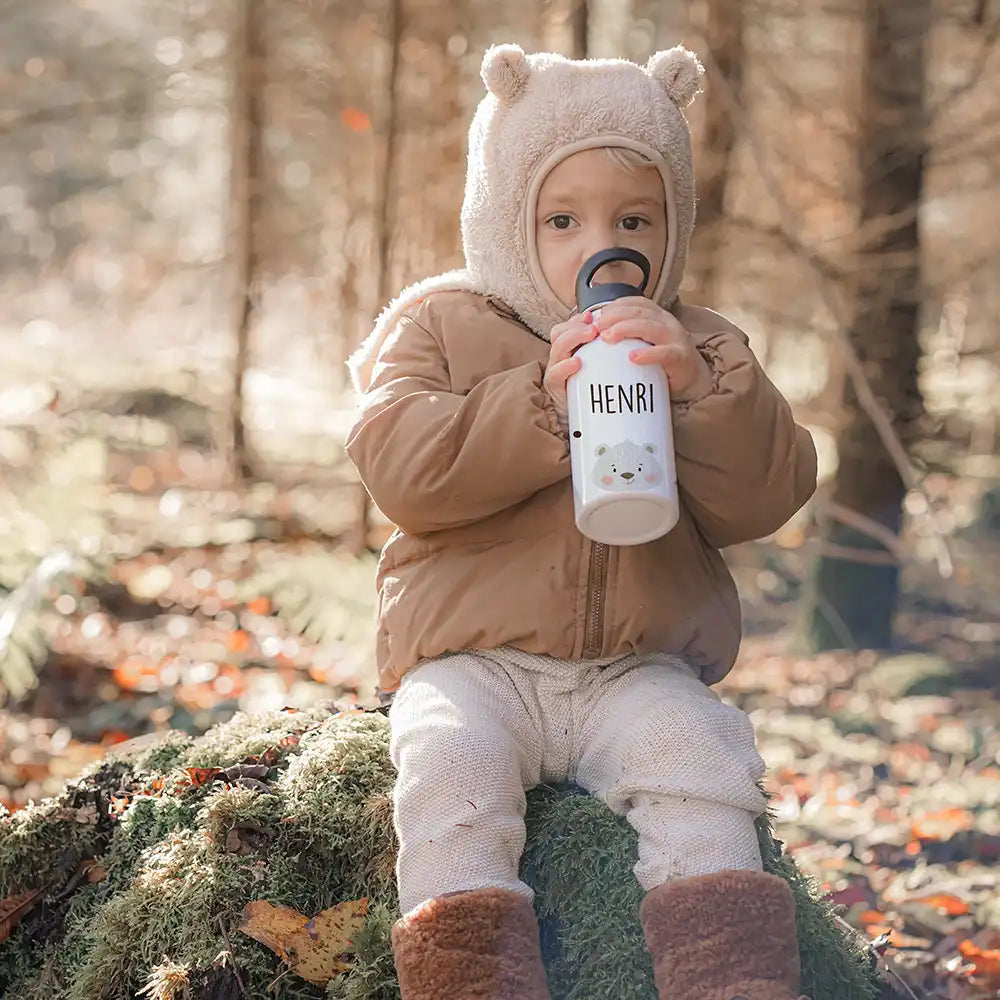 Kind im Wald hält lächelnd personalisierte Kidsmood-Trinkflasche in der Hand - Nahaufnahme eines glücklichen Kindes mit individuell gravierter Edelstahl-Trinkflasche während eines Naturausflugs zwischen Bäumen und grüner Vegetation
