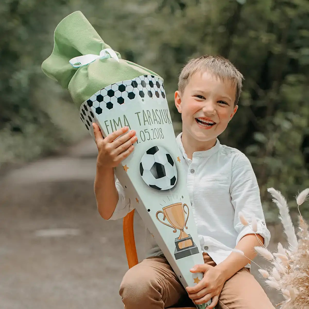 Lachender Junge mit personalisierter Fußball-Schultüte zur Einschulung - stolzer Erstklässler präsentiert seine individuell gestaltete Zuckertüte mit grüner Schleife