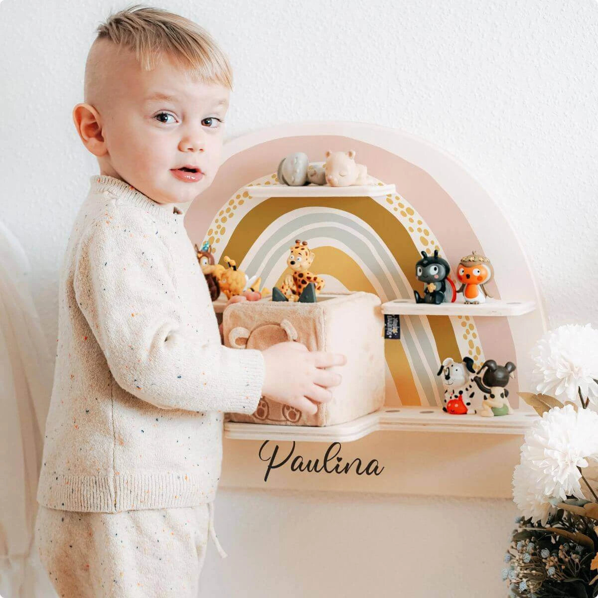 Blondes Kleinkind in cremefarbenem Pullover betrachtet personalisiertes Tonie-Regal mit Regenbogendesign und Namen 'Paulina'. Holzregal mit niedlichen Tierfiguren in pastelligen Farben als stilvolle Aufbewahrungslösung fürs Kinderzimmer