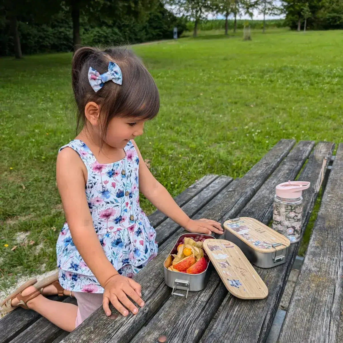 Mädchen mit personalisierter Lunchbox und Trinkflasche beim Picknick - fröhliches Kind im sommerlichen Blumenkleid mit passendem Haarschmuck öffnet ihre individuelle Brotdose mit gesundem Snack im Grünen